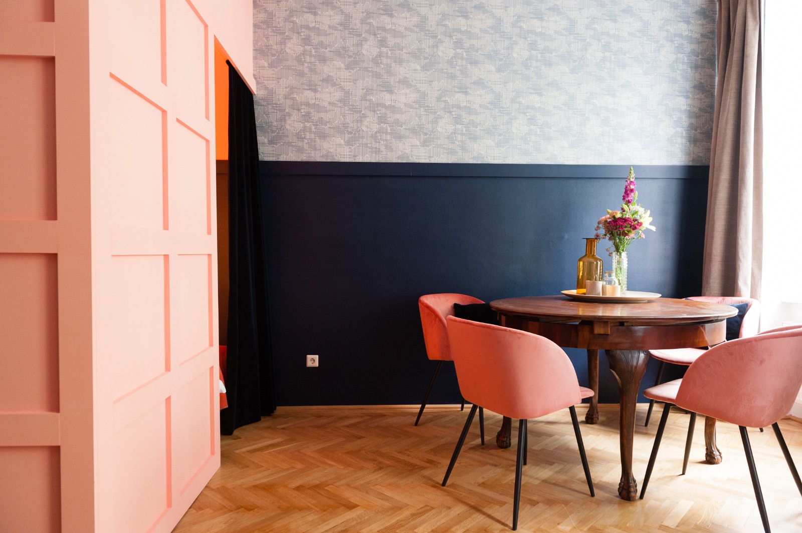 Salon dans un immeuble ancien viennois avec parquet à chevrons, mur bleu, valise vintage en cuir en guise de table, canapé couleur naturelle aux coussins corail et armoire sculptée en noyer