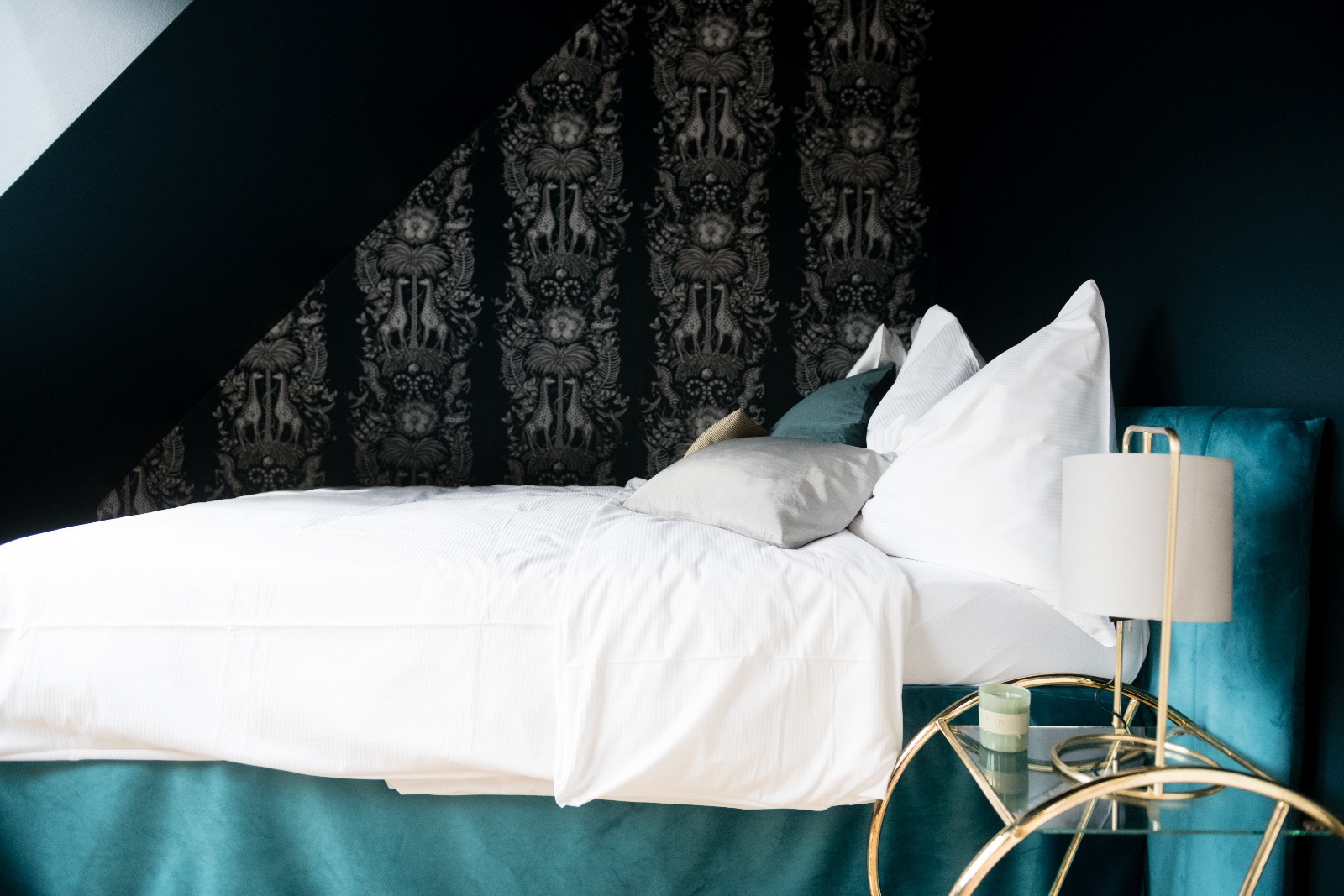 Chambre bleu nuit sous la pente du toit avec tête de lit en velours pétrole, linge de lit blanc et chariot de chevet doré avec lampe et bougie