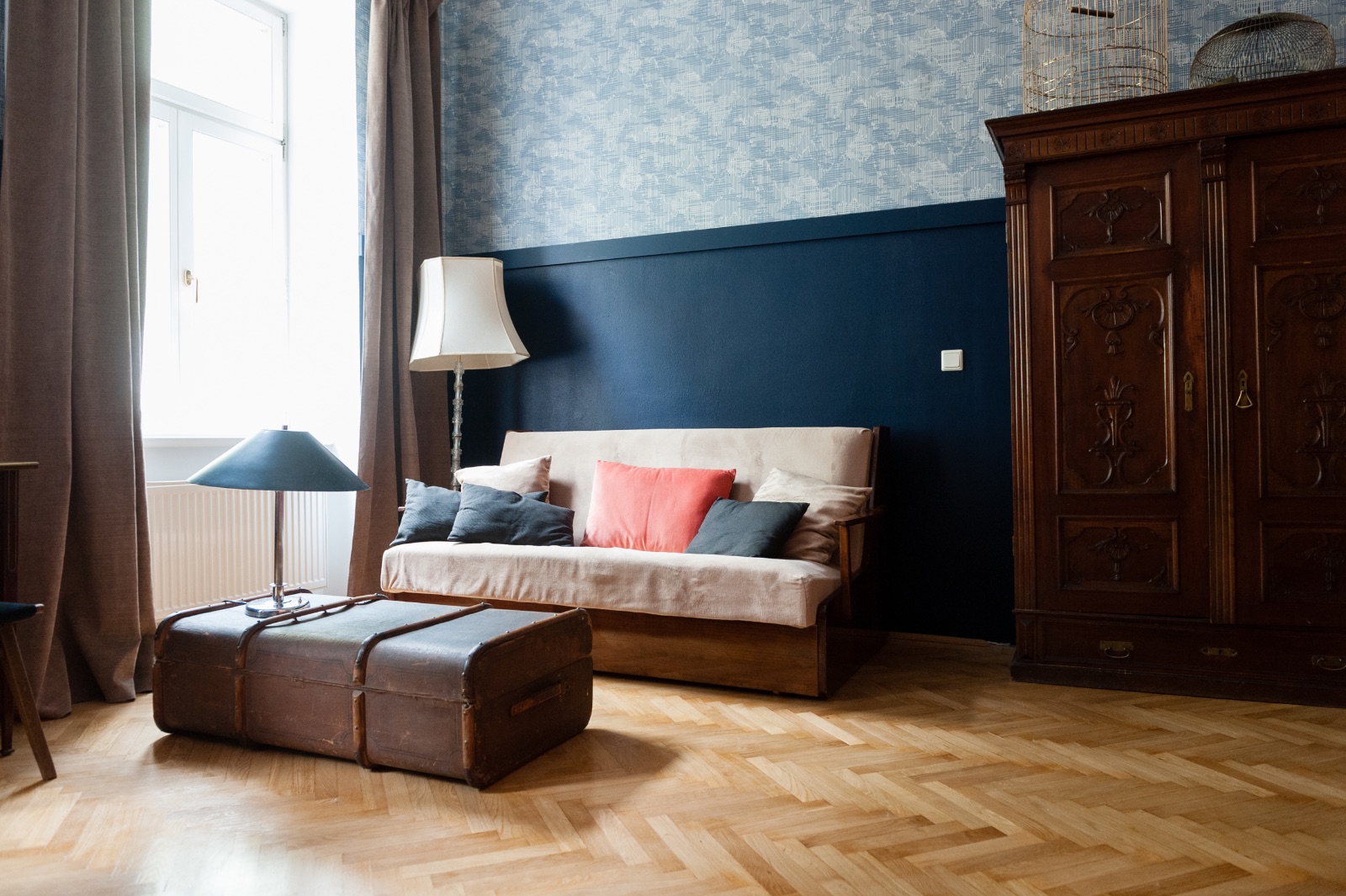 Living room in a Viennese old building with herringbone parquet, a blue wall, a vintage leather suitcase as a table, a natural toned sofa with coral pillows and a carved walnut cabinet