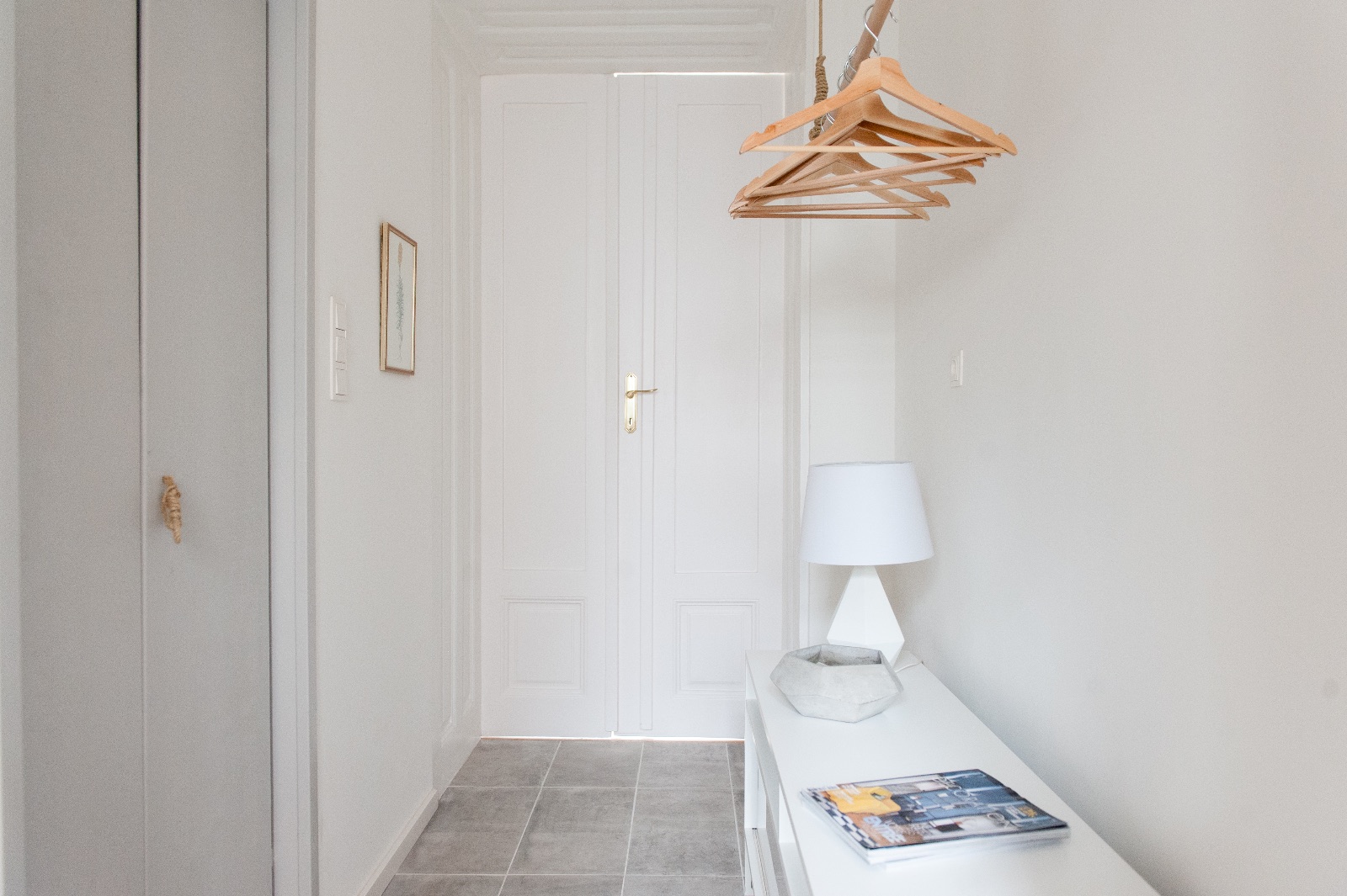 Entrée claire d'immeuble ancien avec cintres en bois suspendus, lampe de table au socle en béton et petit coquillage sur une armoire blanche